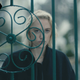 A woman with light blonde hair, wearing a black jacket, stares through an ornate fence in a scene from Watcher.